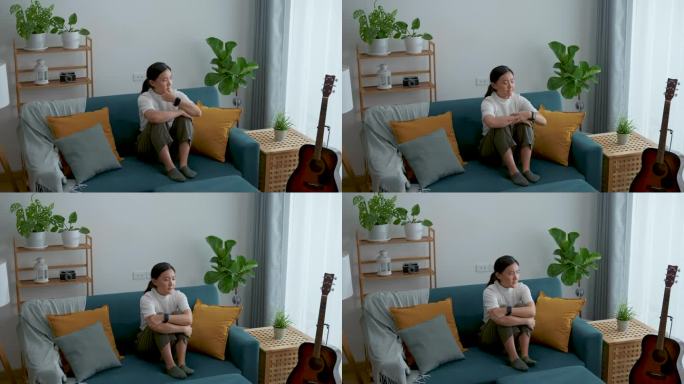 亚洲女人抱着膝盖坐在沙发上，想着悲伤的问题。