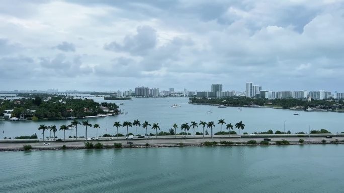 在一个阴天，佛罗里达州迈阿密一条繁忙的高速公路被水包围，远处有热带棕榈树、船只和摩天大楼