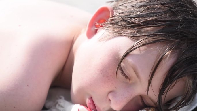 一张10岁男孩的脸，头发湿漉漉的躺在沙滩上。孩子躺着，在阳光下晒日光浴。