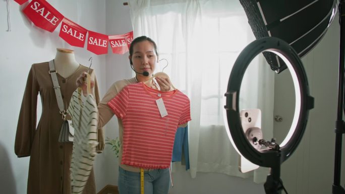 亚洲女性对着智能手机的摄像头在家里卖衣服