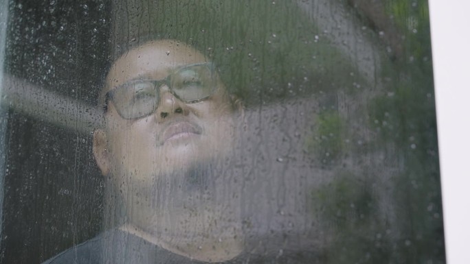 一个身材魁梧的亚洲男人，心情不好，在雨天站在窗前望着窗外，玻璃上的倒影和雨滴。