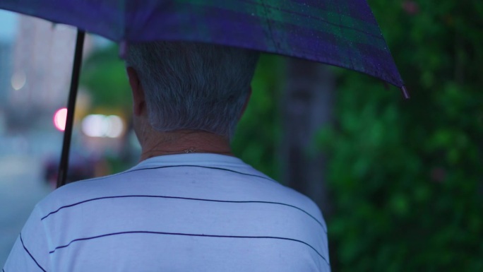 老人在雨天撑伞走在街上的背影。在下雨的下午，在城市环境中散步的老年人
