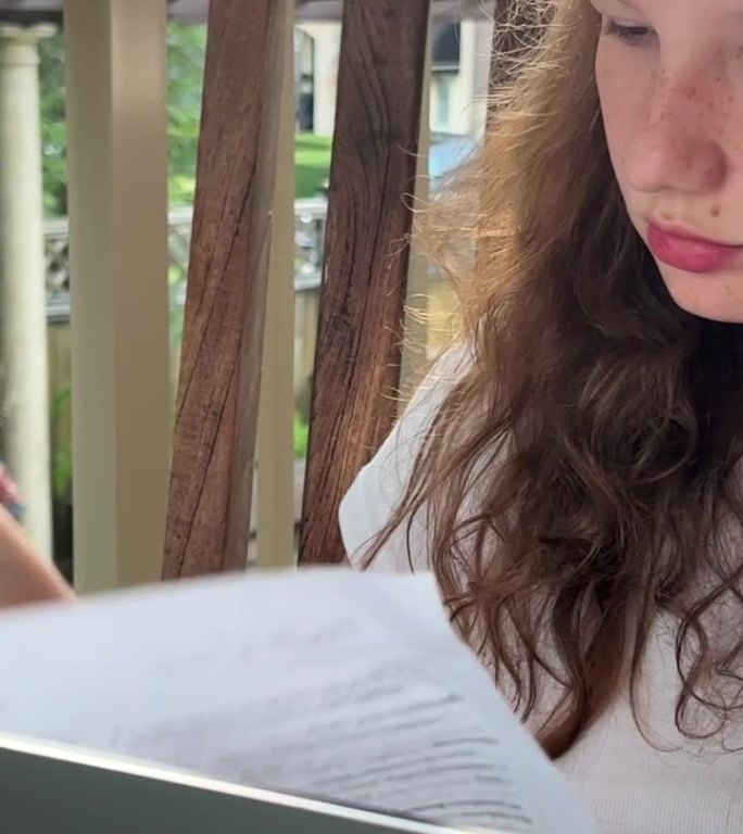 网上学习金发欧洲女孩坐在她的膝上与笔记本电脑在阳台上的街道或观看课程。她的手旁边是打印出来的表格，她