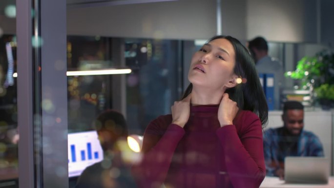 疲惫的亚洲女人按摩脖子，透过摩天大楼的窗户欣赏夜景