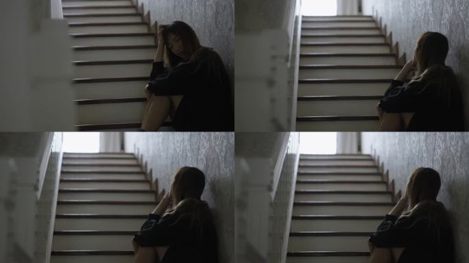 伤心欲绝的亚洲年轻女子，双膝翘起，双手托着下巴，坐在楼梯脚下，茫然地望着楼上，感到沮丧和遗憾。