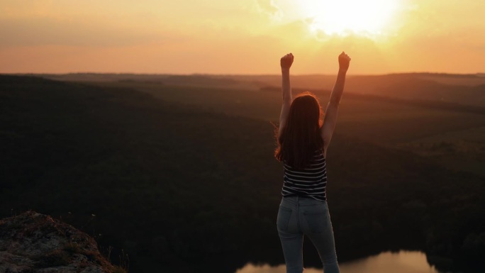 快乐苗条的女孩站在夕阳山，举起双臂，欣赏风景