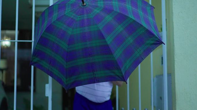 老人撑开伞挡雨，从家里走到街上。老人离开住所，在下雨时打开大门在城市人行道上行走