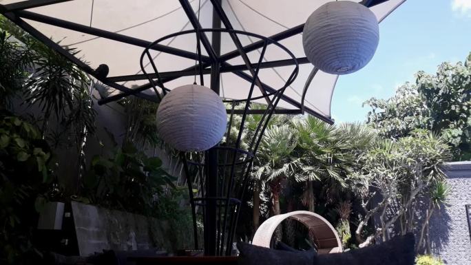 咖啡厅里的白色灯笼装饰灯下有一把大伞，桌椅周围有观赏植物