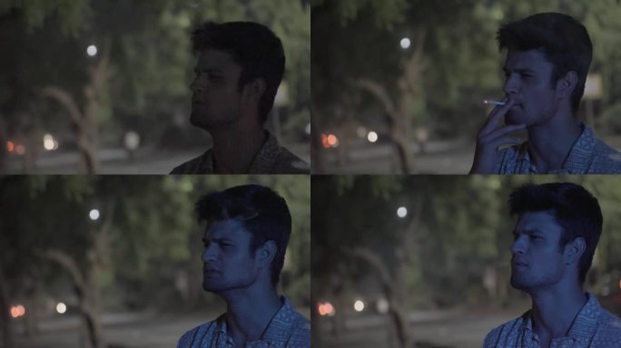 在假期的晚上，一名年轻的印度男子站在树旁抽烟，灯光落在他身上
