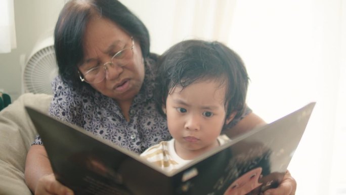 一位亚洲老妇人正坐在沙发上看书。可爱的小男孩投入祖母的怀抱，兴奋地读着书中的故事。