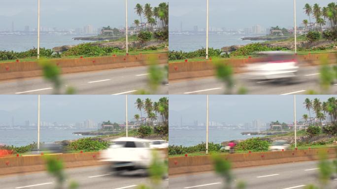 在模糊的前景中，许多汽车和卡车在高速公路上快速行驶。背景是棕榈树和圣多明各市中心。大城市的生活