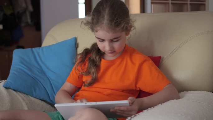 可爱的女孩在平板电脑上玩游戏的肖像，在客厅放松。孩子浏览社交媒体。孩子坐在沙发上使用平板电脑，在家上