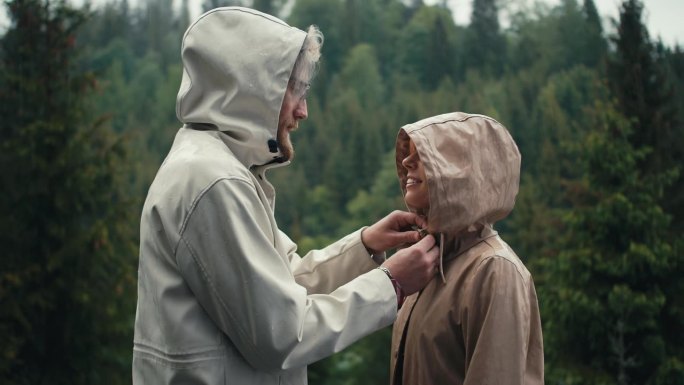 在山间针叶林的雨中，男孩系好女孩的外套，吻了她。绿色森林背景下的幸福情侣