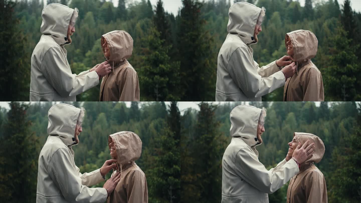 在山间针叶林的雨中，男孩系好女孩的外套，吻了她。绿色森林背景下的幸福情侣