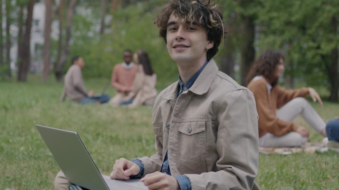 一个微笑的年轻人坐在公园的草坪上，拿着笔记本电脑看着镜头