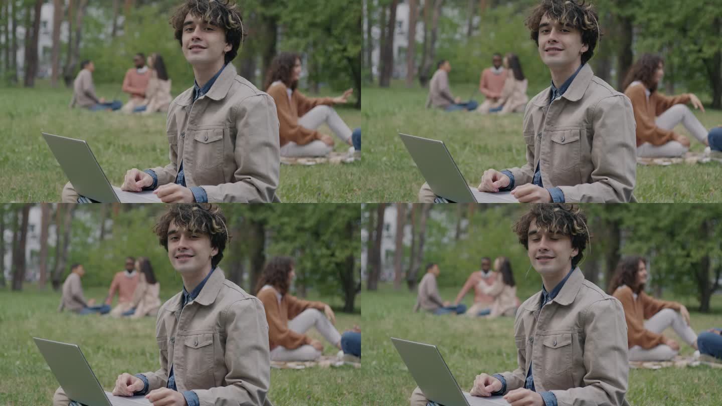 一个微笑的年轻人坐在公园的草坪上，拿着笔记本电脑看着镜头