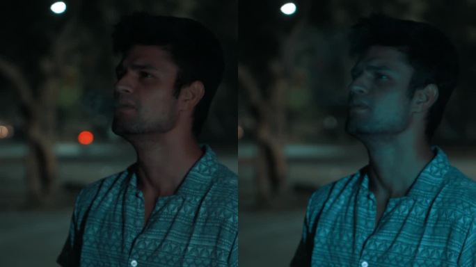 在假期的晚上，一名年轻的印度男子站在树旁抽烟，灯光落在他身上
