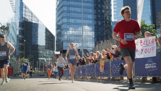 一群不同的人在城市马拉松比赛中竞争的慢动作地面镜头。观众中的家人和朋友热情地欢呼并支持他们所爱的人