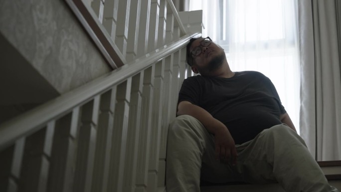一个身材高大的亚洲人，在失去工作后，疲惫地坐在家里的楼梯上，仰望着空虚的眼睛，感到绝望。