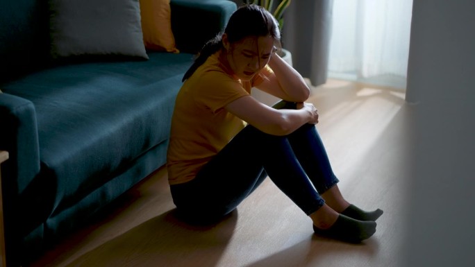 亚洲女人抱着膝盖坐在客厅里，想着问题感到悲伤。