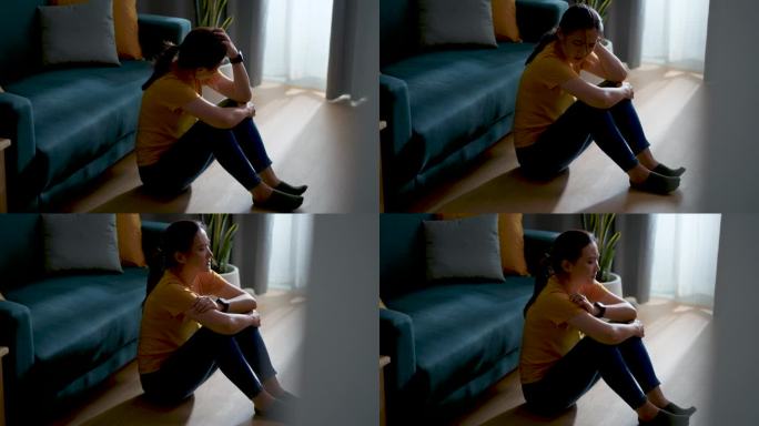 亚洲女人抱着膝盖坐在客厅里，想着问题感到悲伤。