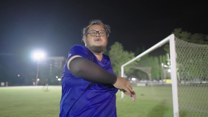 一个疲惫的超重的亚洲男人站在足球场上做三头肌伸展，上半身摆动，手臂向前滚动，胸部和肩膀伸展，以提高身