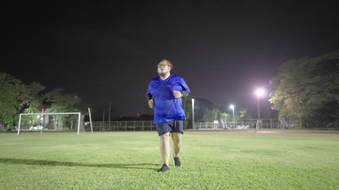 为了减肥，一个非常胖的亚洲男人在公园的草坪上继续他的跑步运动，尽管已经很晚了。