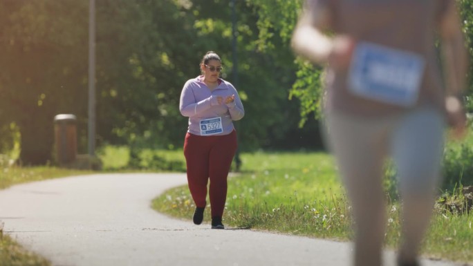 慢动作肖像一个加码的女性马拉松运动员在比赛期间休息和散步在一个绿色公园。女性慢跑者感到不舒服，疲倦，
