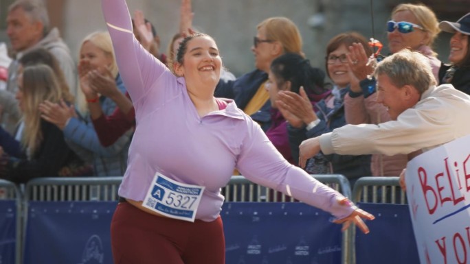 一个微笑的加码女运动员穿过终点线并展示她的意志力的慢动作肖像。友好的城市马拉松观众支持并与参赛者击掌