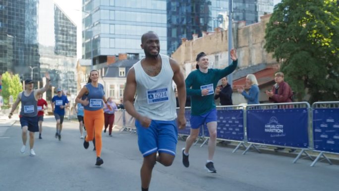 一个微笑的黑人青年在城市马拉松比赛中向支持他的观众挥手的慢动作肖像。友好快乐的男运动员庆祝在比赛中越