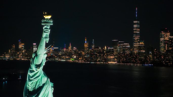 电影直升机在夜晚经过自由女神像。被照亮的罗马女神手持火炬的近距离全景航拍镜头。地标性的纽约纪念碑和曼