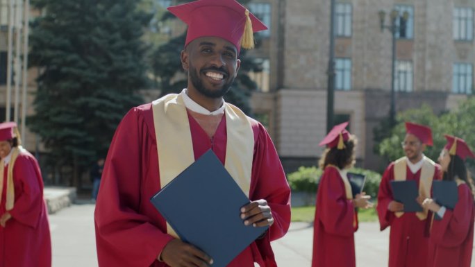 一个成功的非裔美国人的慢动作肖像，他穿着毕业袍，戴着毕业帽，站在校园里拿着毕业证书