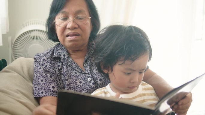 亚洲小孙子试图在家里和慈爱的奶奶一起读书。蹒跚学步的男孩喜欢和奶奶讲有趣的故事。