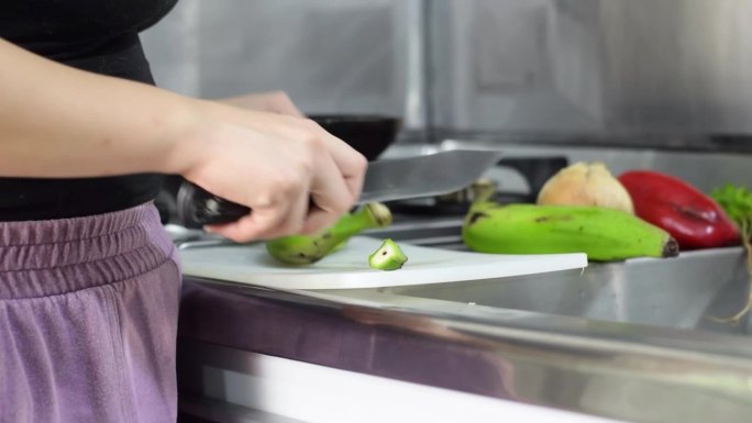 一名妇女在厨房用刀切绿香蕉准备食物的详细视图。