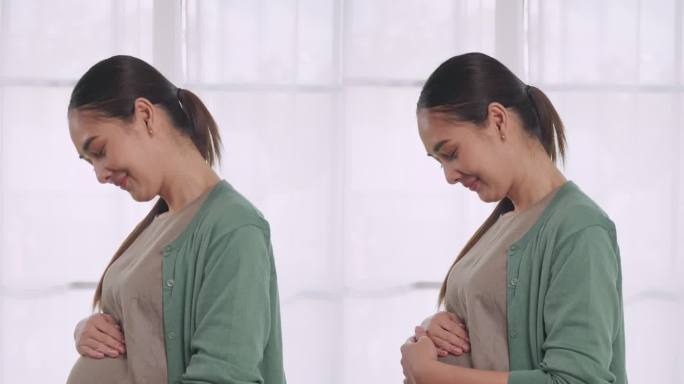 垂直夹。快乐的亚洲孕妇站在房子的窗户边，深情地抚摸着她未出生的孩子。