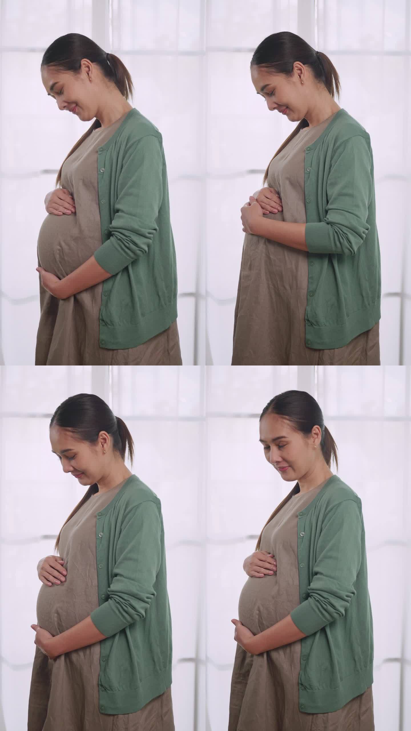 垂直夹。快乐的亚洲孕妇站在房子的窗户边，深情地抚摸着她未出生的孩子。