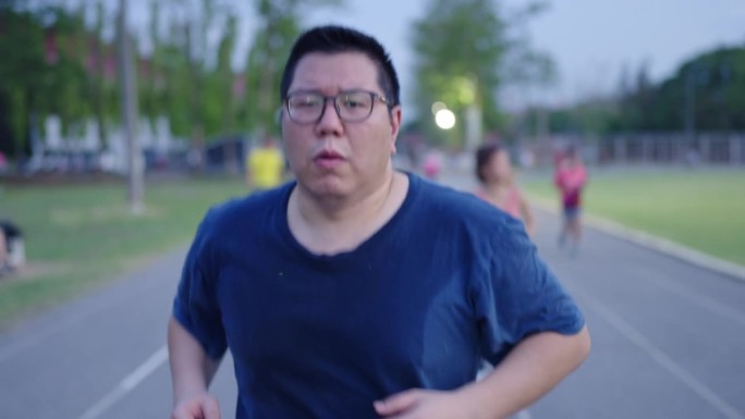一名肥胖的亚洲运动员在体育馆的跑道上气喘吁吁地跑着，带着疲惫的表情看着镜头，背景是其他在跑道上跑步的