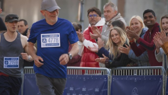 一名老年男性慢跑者在城市马拉松比赛中奔跑并受到观众欢呼的慢动作肖像。健康健美的老人享受体育活动，保持