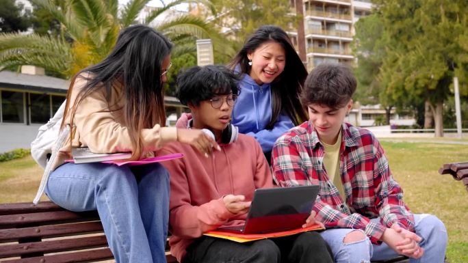 回到学校。一群多民族青少年使用笔记本电脑坐在高中外的长凳上。