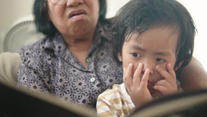 亚洲细心的孙子坐在沙发上听奶奶读童话书。