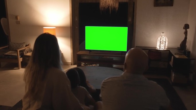 一家人带着孩子在家看电视。父母和孩子在晚上客厅看电视屏幕模拟的白色色彩。休闲人士和小孩的休息时间