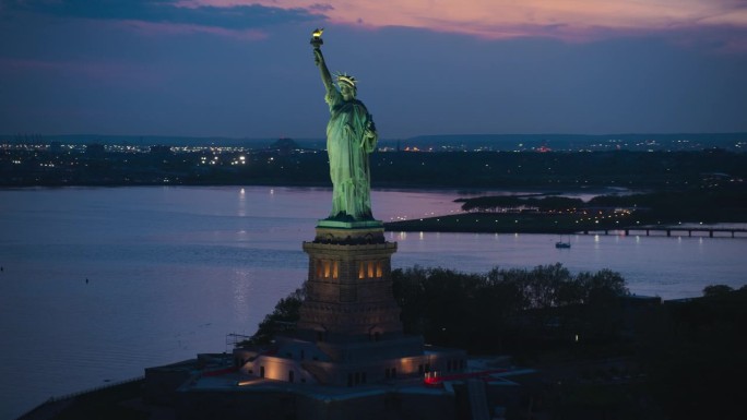 自由女神像周围的空中飞行。直升机拍摄的爱国纪念碑在美丽的日落背景下，站在纽约市港口的自由岛上