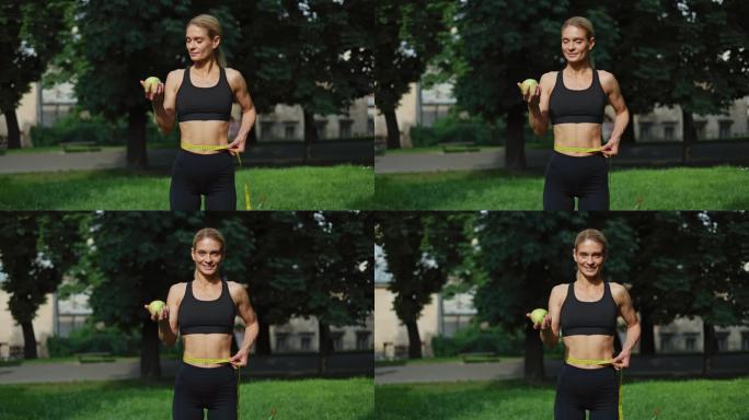 在新鲜空气中用黄色厘米测量腰部体积的苗条高加索女运动员的正面视图。活泼迷人的女性拿着健康的绿色水果，