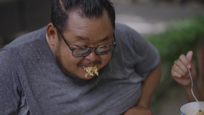 延时:在公园野餐时，一个饥饿的肥胖男子用叉子嚼着一口意大利面，和他的朋友们聊得很开心。