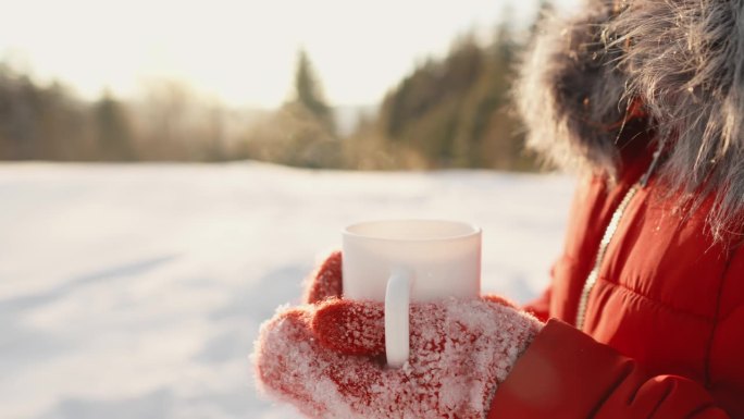 冬天的院子里，一个女人拿着一个白色的杯子，里面装着热气腾腾的热饮。美味的茶或芳香的咖啡。雪里的热葡萄