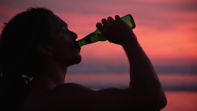 男子在海边或海水的夏日日落边喝啤酒。现代人在假期放松时喝一杯冷饮。在温暖的自然景观的浪漫光线下，热辣