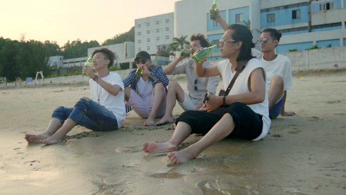 年轻的亚洲成年人坐在海滩上放松, 喝啤酒敬酒.