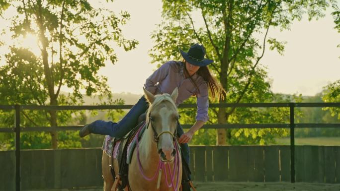 戴着牛仔帽的年轻女子在被树木环绕的牧场上从马背上下来
