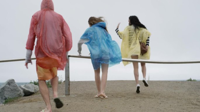 在雨天的海滩上，一群十几岁的朋友穿着五颜六色的雨衣雨披爬过栏杆跑慢动作，庆祝冒险