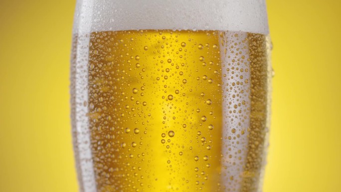 摄像机沿着装有淡啤酒的啤酒杯缓缓升起。玻璃上有很多水珠，上面还有啤酒泡沫。黄色背景。